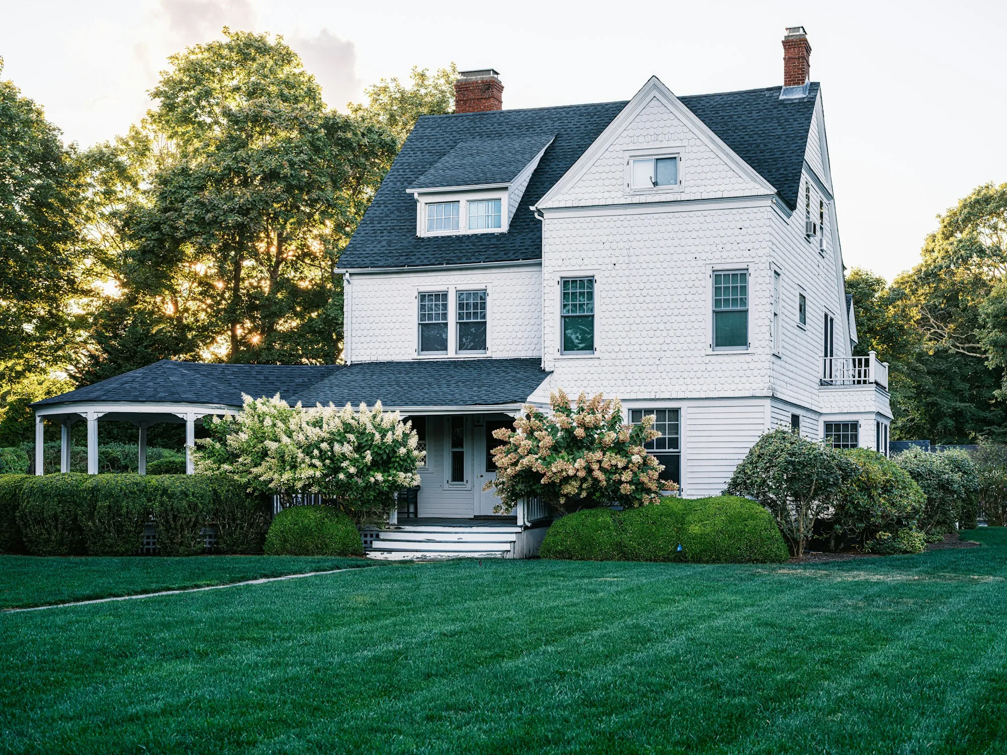 White shingle style house with dormers under a dark asphalt roof, seen across a clipped hedge